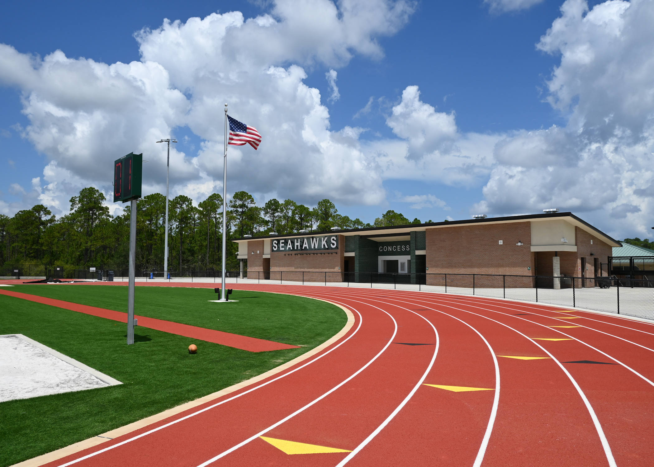 A view of the bright red running track curving around a green artificial turf field with an American flag on a tall pole. In the background is the concessions building with the large word "SEAHAWKS" displayed prominently.